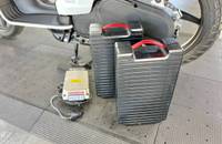 Ein weißer Elektroroller mit mehreren extern platzierten Akkus und einem Ladegerät.