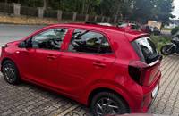 Roter KIA Picanto von hinten links auf einem Pflastersteinparkplatz vor einem Autohaus.