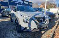 Außenansicht eines grauen Nissan Juke, geparkt vor einem Autohaus mit blauem Gebäude.
