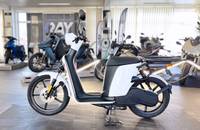 Seitenansicht eines weißen Elektrorollers (E-Scooter) mit schwarzem Akku und Sitz, abgestellt in einem Showroom.