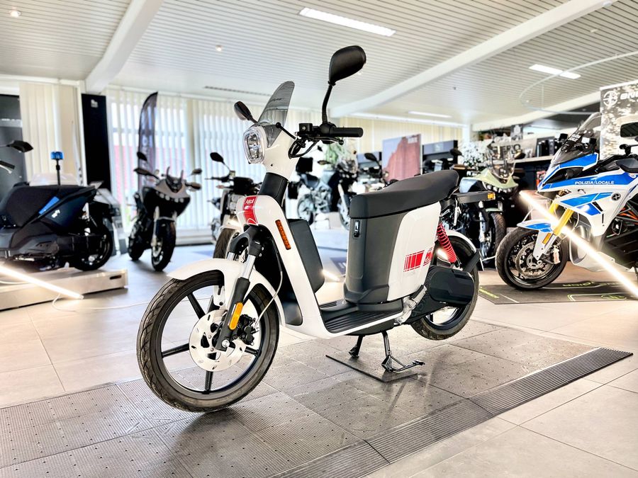 Ein weißer Elektroroller mit schwarzem Akku steht auf einem grauen Boden in einem Showroom.