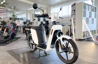 Dreiviertelansicht eines weißen Elektro-Scooters in einem Showroom. Das Fahrzeug steht auf einem Teppichboden.