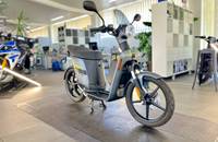 Ein grauer Elektroroller steht in einem Showroom. Man sieht die Seiten- und Frontansicht des Fahrzeugs.