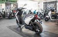 Ein weißer Elektroroller steht in einem Showroom mit weiteren Motorrädern im Hintergrund.