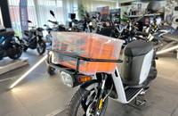 Außenansicht eines weißen E-Scooters in einem Showroom. Das Fahrzeug ist mit einem orangefarbenen Frontgepäckträger ausg