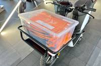 Ein weißer Elektroroller mit einem transparenten Kasten auf dem Heckträger. Der Kasten enthält orangefarbene Dokumente o