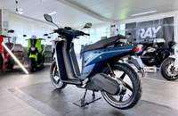 Seitenansicht eines blauen Elektrorollers (Scooter) in einem hellen Showroom. Das Fahrzeug steht auf einem grauen Boden.