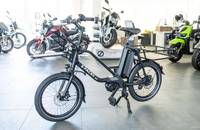 Schwarzes Lasten-E-Bike (E-PACKR 8.0) in einem Showroom, umgeben von anderen E-Bikes.