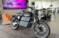 Schwarzes Elektro-Motorrad in einem Showroom. Im Hintergrund sind weitere Fahrzeuge und ein Bildschirm zu sehen.