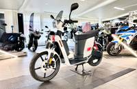 Ein weißer Elektroroller steht in einem hellen Showroom, umgeben von anderen Fahrzeugen.